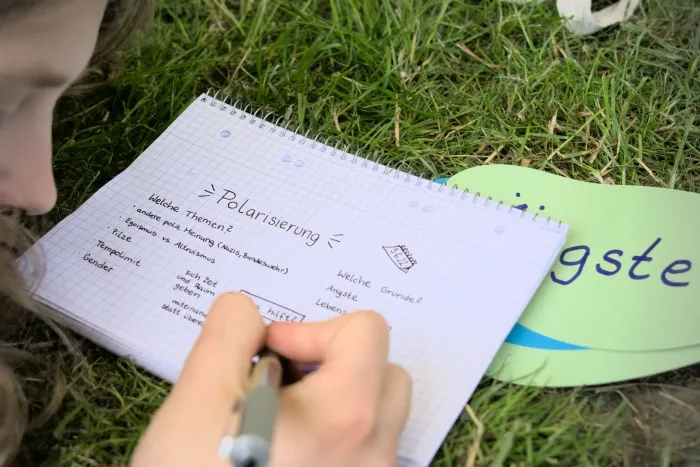 Blick über die Schulter einer Person, welche auf dem Rasen liegt und auf einem Blatt Papier Notizen zum Thema 'Polarisierung' verfasst.